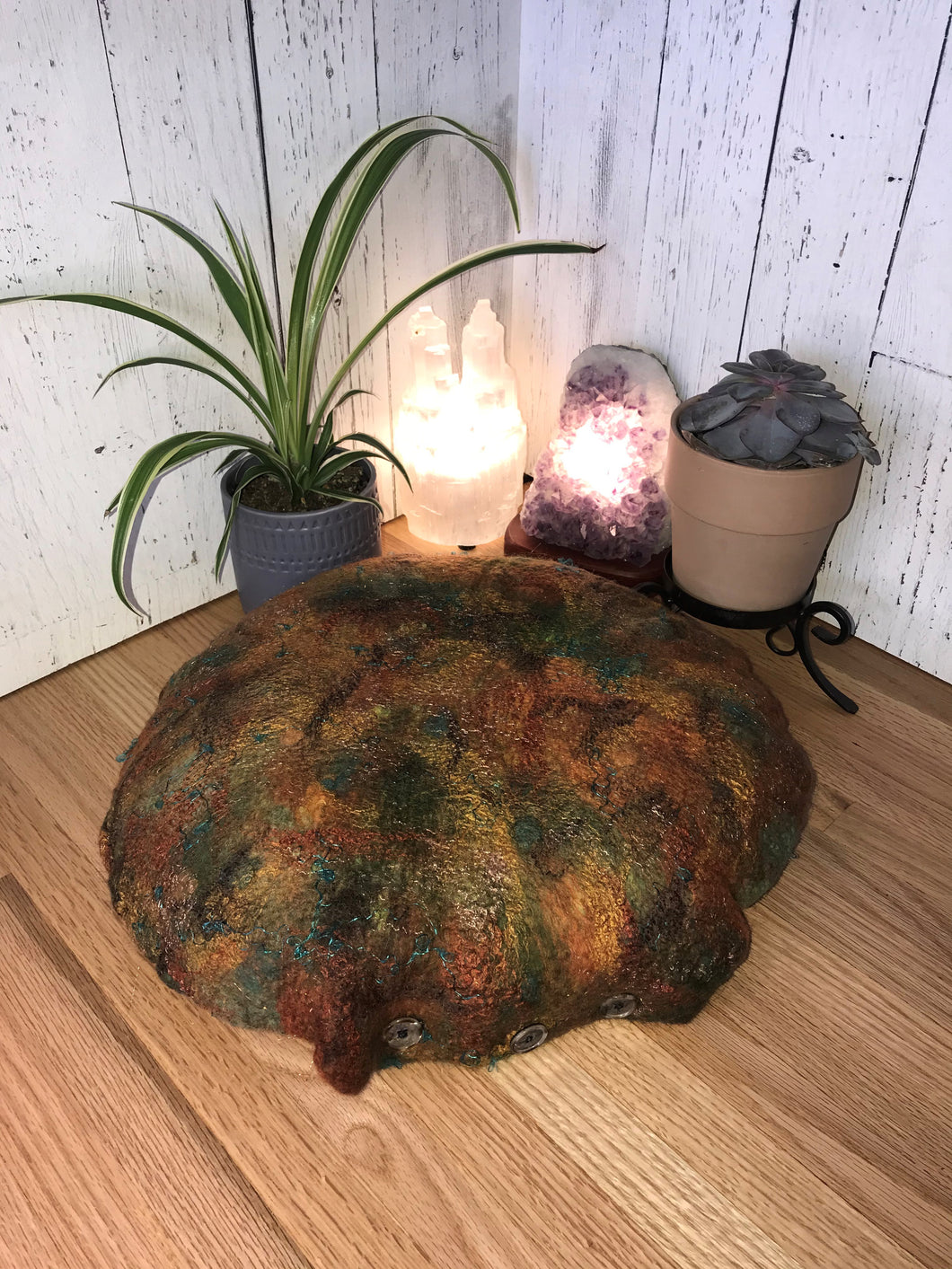 lifestyle photo of  round meditation floor cushion felted with merino wool into silk fabric with tussah silk bamboo accents mixtures of various shades of green, rust, and patina, cooper and teal shimmery accents. There is a small spider plant a succulent and a selenite, amethyst crystal lamp in the background
