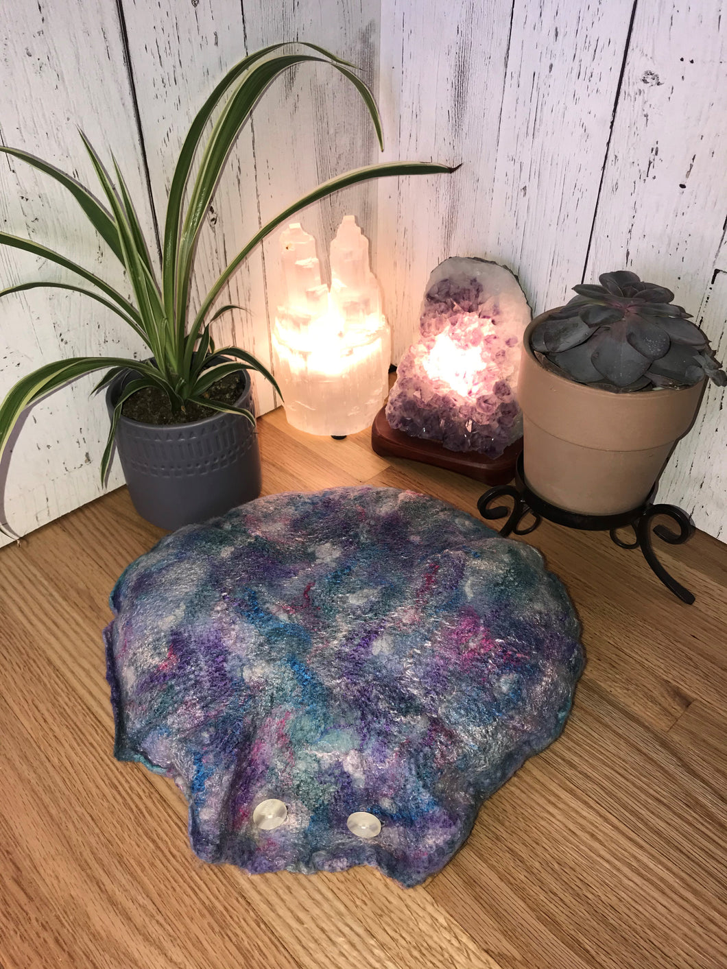lifestyle photo of a round meditation flor cushion made of felted soft merino wool into silk fabric with silk and bamboo accents for a shimmery luster various shades of blues purple and teal with hints of pink and white small green spider plant and succulent with a selenite crystal tower lamp and amethyst crystal lamp in the background
