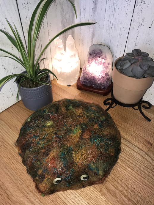 lifestyle photo of an artisan meditation floor cushion made from soft felted merino wool into silk fabric with silk, bamboo, and other natural fiber accents for a pop of color and shimmer. various shades of browns and greens mixed with rust and patina with hints of teal and copper. A small spider plant and succulent plant with a selenite tower lamp and an amethyst crystal lamp in the background
