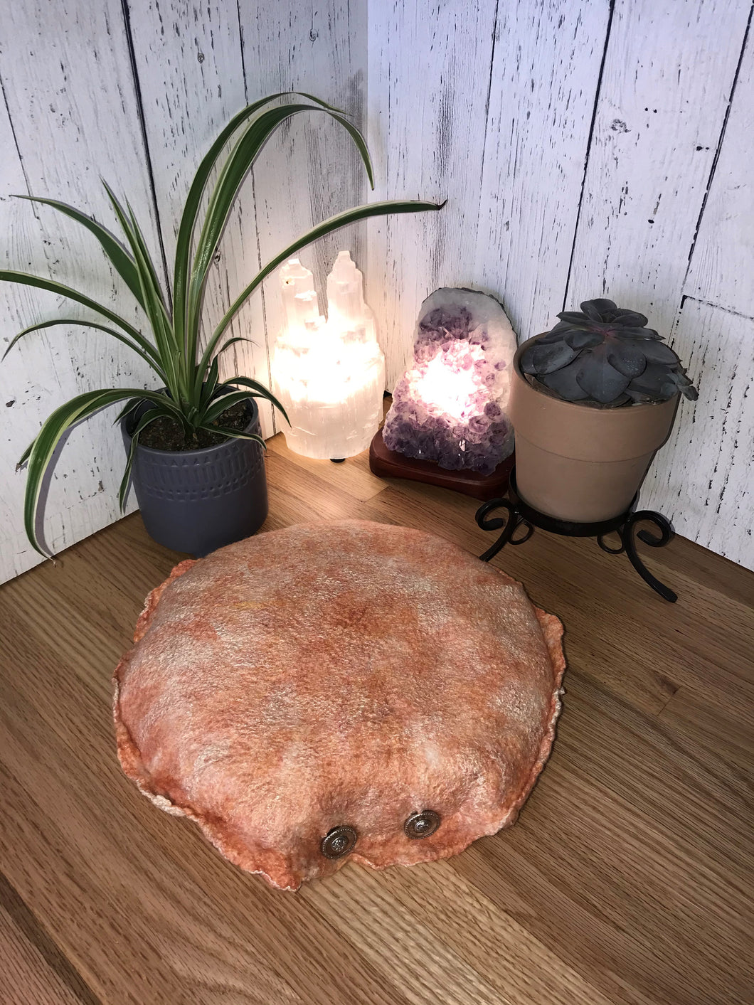 lifestyle photo of a round meditation floor cushion made from felted soft merino wool into silk fabric with silk accents to give the cream and peach wool a soft shimmer. A small spider plant and succulent is in the background along with a selenite tower lamp and an amethyst crystal lamp.