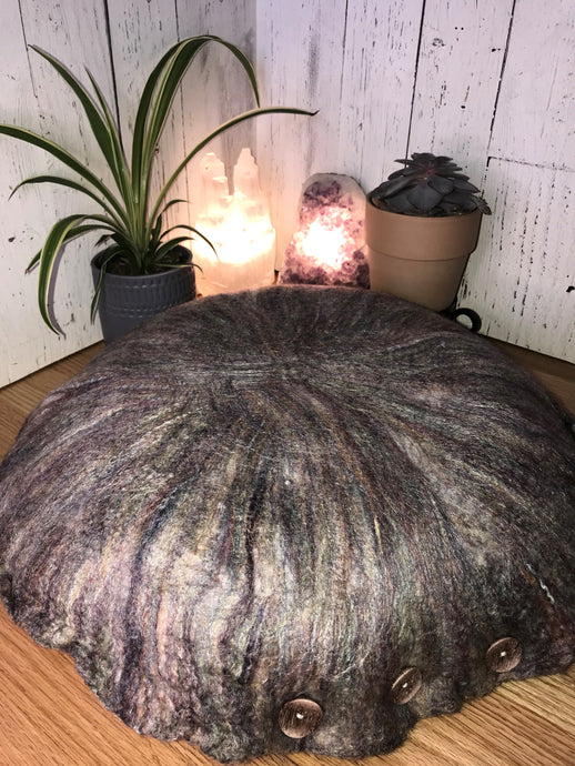 lifestyle phot of round artisan floor meditation cushion hand felted with a soft merino wool into silk fabric rose colors dominate however there is  hints of burgundy, purple, mint green, green yellow and cream colors. A small plant and succulent along with a white selenite tower lamp and an amethyst crystal lamp in the backgound
