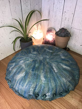Load image into Gallery viewer, lifestyle photo of artisan floor meditation cushion made with luxurious soft merino wool felted into silk fabric with silk accents the blues and greens mix with white shimmers of tussah silk as an accent. A small plant along with a succulent and a white selenite tower lamp and a amethyst crystal lamp in the background
