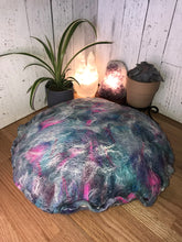 Load image into Gallery viewer, Bottom view of lifestyle phot of &quot; Unicorn&#39;s Breath Meditation Cushion&quot; a round artisan made floor meditation cushion. hand made with merino wool felted into silk fabric with a leopard print various shades of blues teals some purples pinks and white shimmer. A small green plan a succulent plant white selenite tower lamp along with a amethyst crystal lamp in the background
