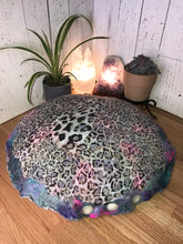 Load image into Gallery viewer, lifestyle phot of &quot; Unicorn&#39;s Breath Meditation Cushion&quot; a round artisan made floor meditation cushion. hand made with merino wool felted into silk fabric with a leopard print various shades of blues teals some purples pinks and white shimmer. A small green plan a succulent plant white selenite tower lamp along with a amethyst crystal lamp in the background
