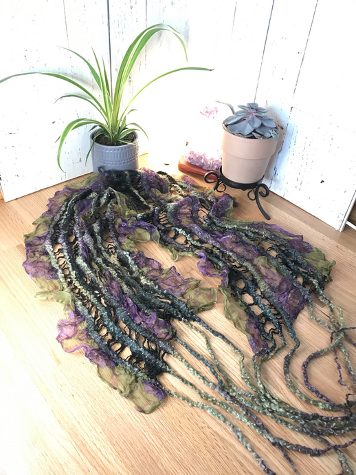 lifestyle product photo of an artisan scarf prayer shawl hand felted from merino wool/ tencel blend and a green and purple floral silk chiffon fabric. the felted wool creates soft ruffles in the silk fabric and tussah silk give a lace vibe to the soft and flowing unique piece. there is a small plant and succulent plant along with a white selenite tower lamp and amethyst crystal lamp in the backgound